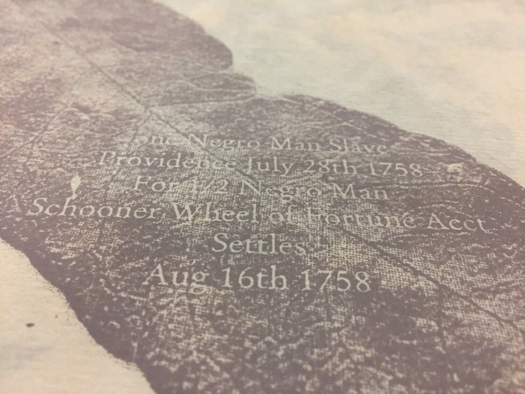 Close-up of a page from "Harvest: Holding & Trading" which shows text knocked out of a brown leaf printed on tan paper. The text is a 1758 listing for a slave.