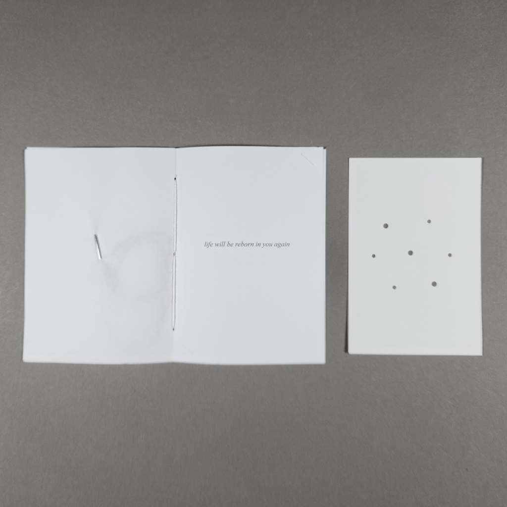 An open pamphlet, with embroidery needle and line of poetry inside, next to a hole-punched card.